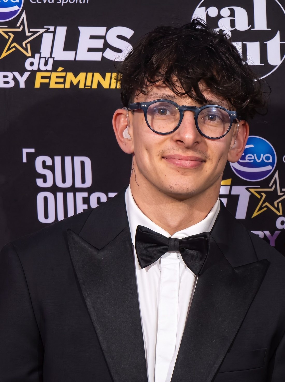 Portrait d’Arthur Rimour, lunettes rondes et nœud papillon, devant le panneau des Étoiles du Rugby Féminin.