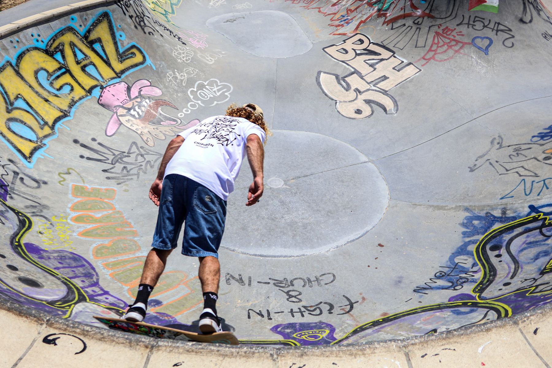 Le modèle en tee-shirt blanc descend un bowl de skatepark couvert de graffitis.