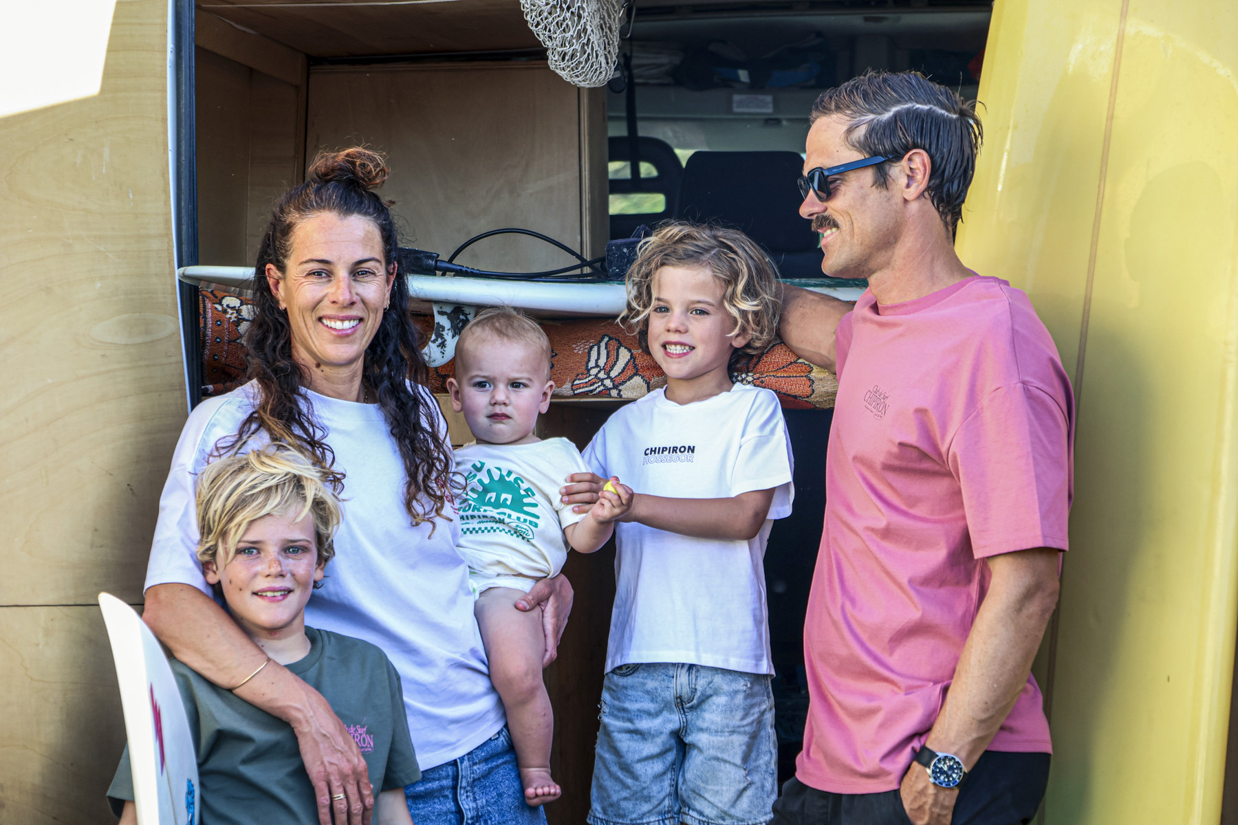 Une famille devant son van ouvert, planches de surf jaunes de chaque côté.